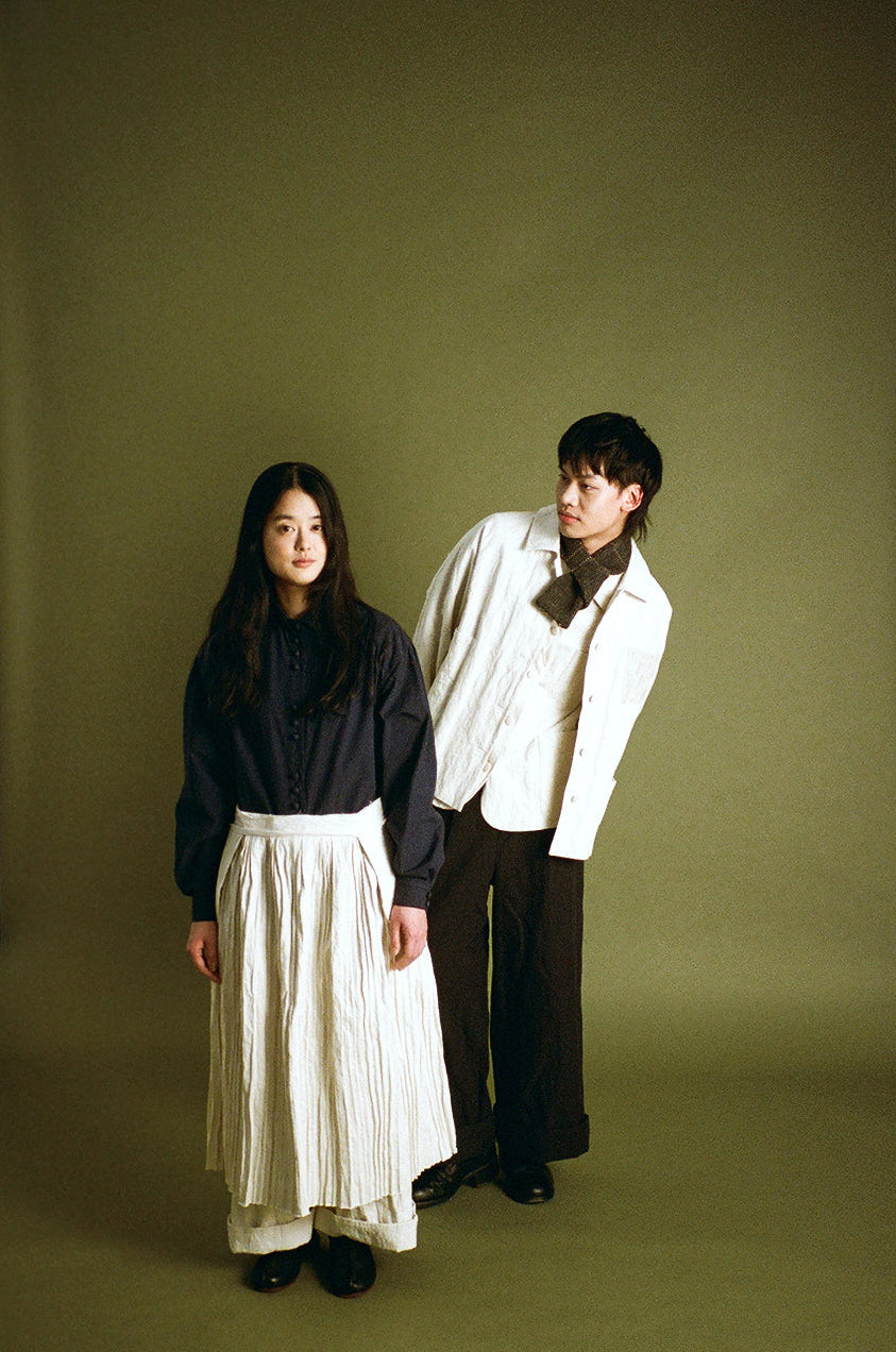 A man and a woman standing in front of an olive green studio backdrop. The woman wears a dark navy long-sleeve top and a cream pleated midi skirt, while the man wears a cream textured jacket, dark trousers, and a dark bow tie.