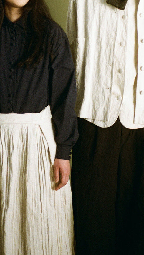 A man and a woman standing side by side against a solid olive green studio background. The woman wears a black long-sleeve top and a long cream pleated skirt.