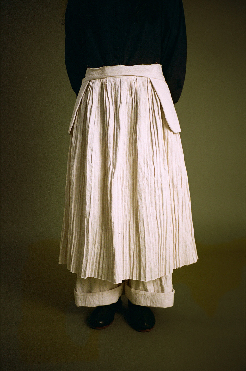 A person shown from the back wearing a long pleated cream skirt with a matching waistband and side ties, paired with a dark top and black shoes, against a dark olive background.