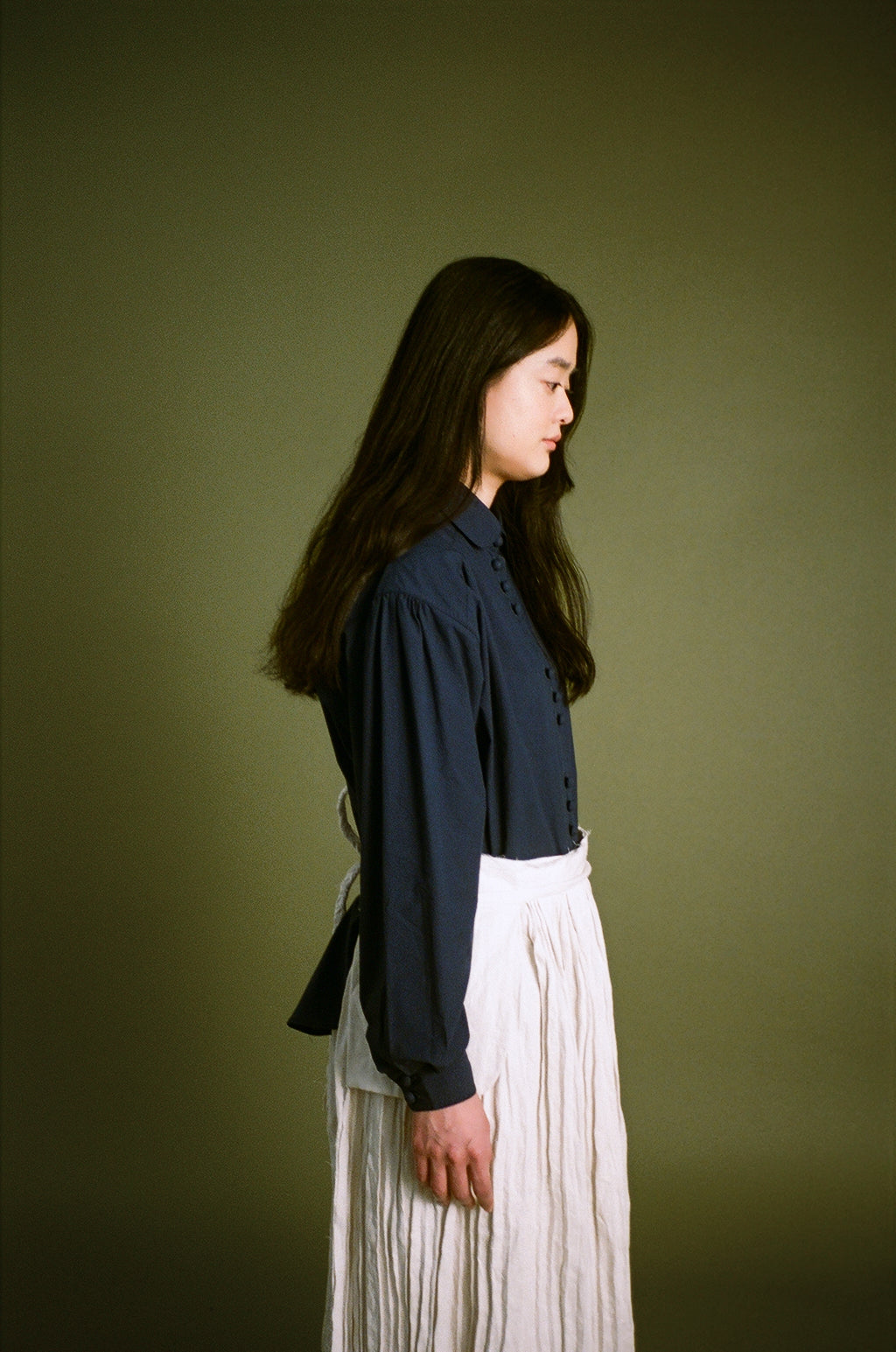 A woman in profile wearing a dark navy long-sleeve button-up shirt and a high-waisted white pleated skirt, standing against a solid olive green studio background.