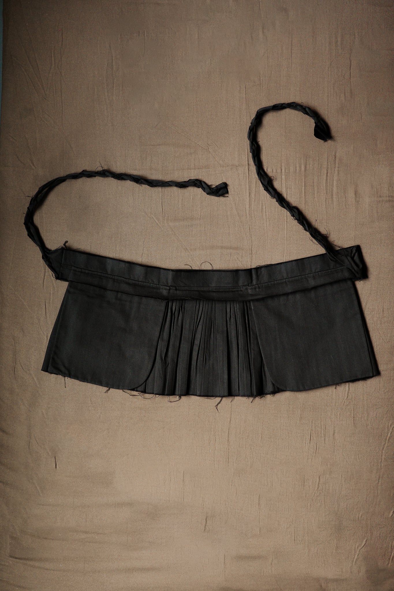 Black pleated Goma apron with a structured waist band and braided ties.