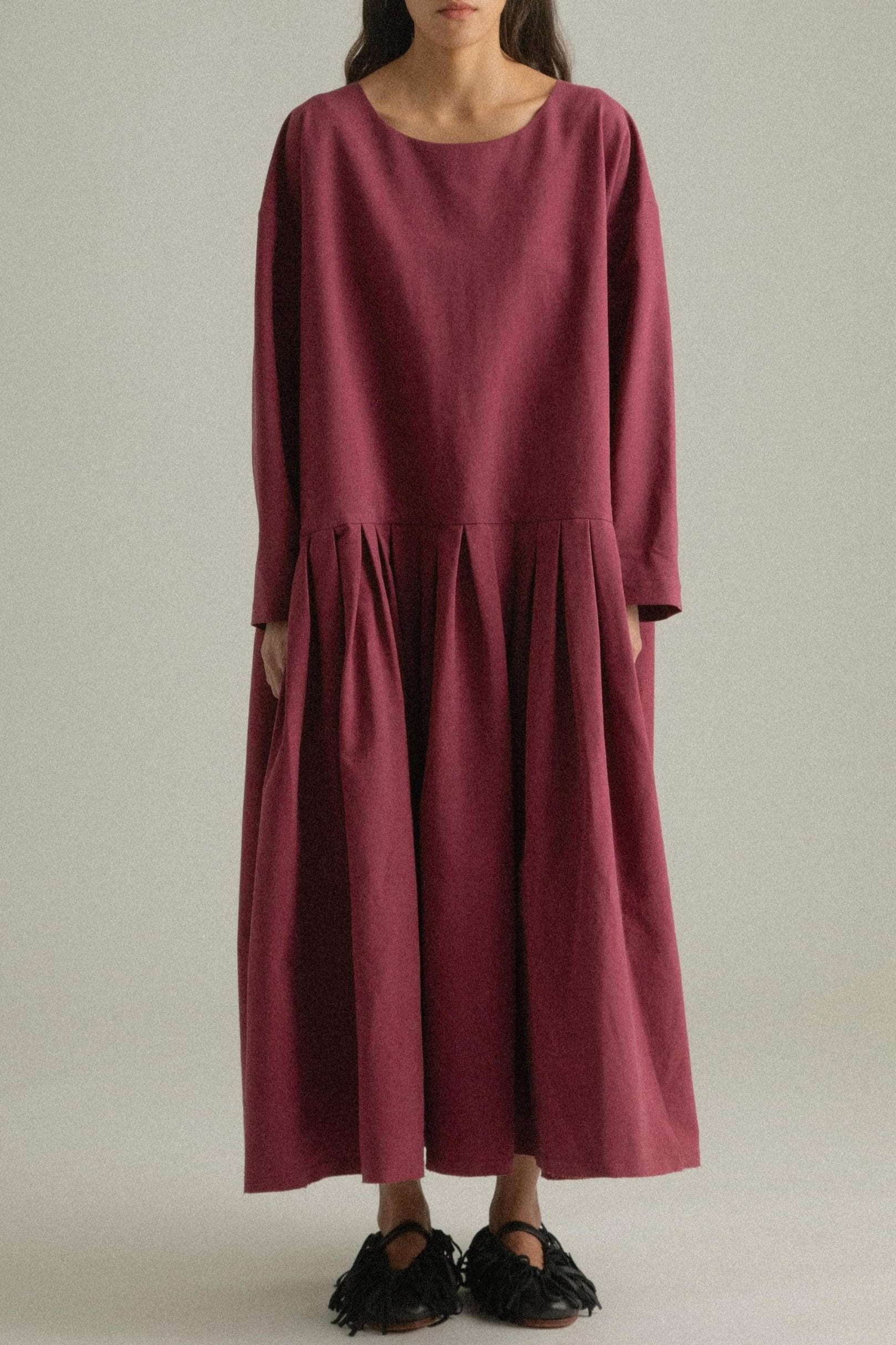 A model wearing the Arya Dress, crafted from 100% bony cotton with a loose, asymmetric round neckline and ankle-length gathered pleats. The dress can be worn front-to-back or back-to-front and features side pockets. The model is shown in front, side, and back views.