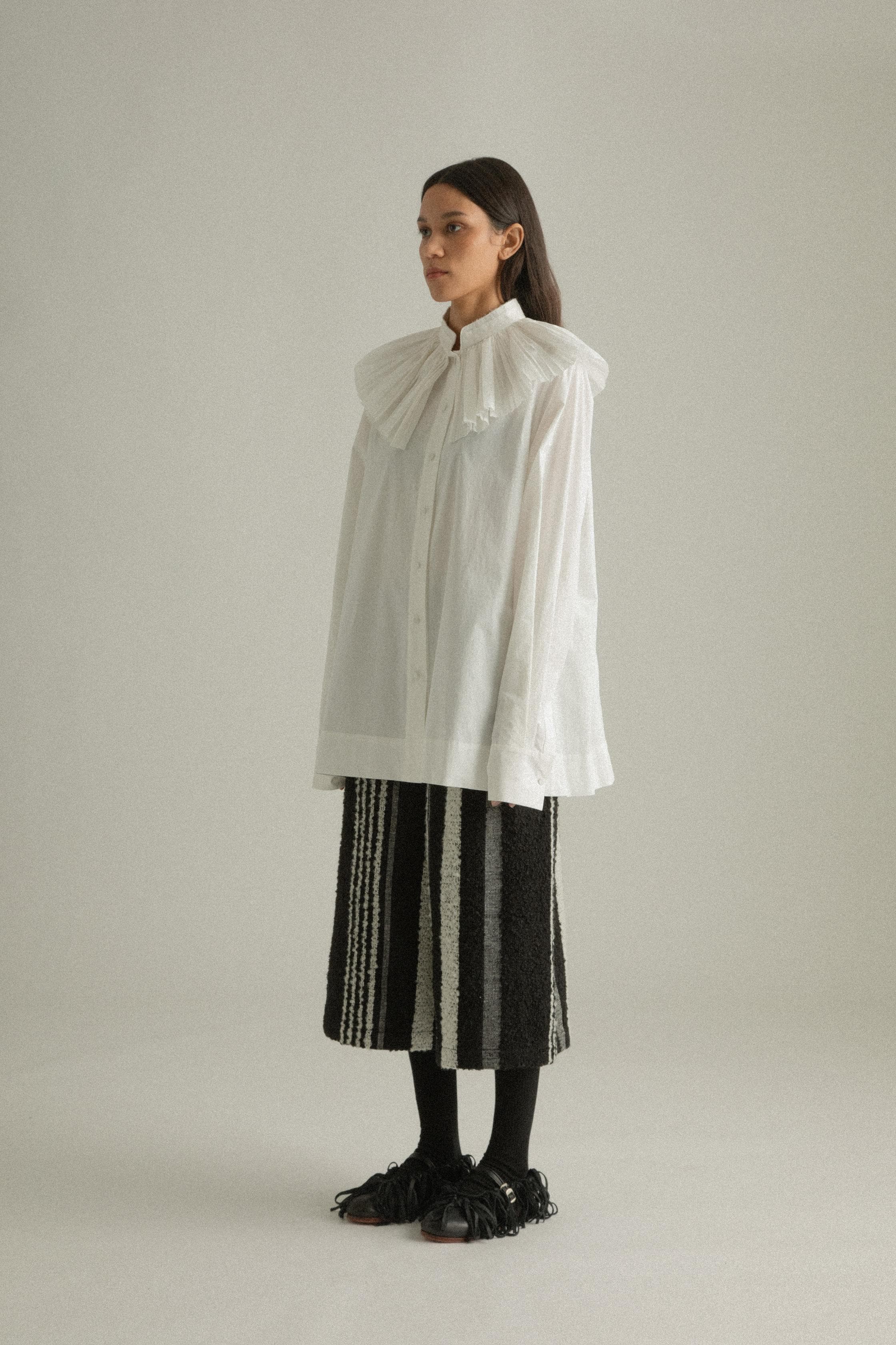 A model wearing the Carcy Shirt, a relaxed-fit top made from 100% organic cotton. It features a large ruffled collar with Chinese-inspired detailing. The shirt is paired with DI Pants to create a layered, textured ensemble. The model is shown in front, side, and back views.