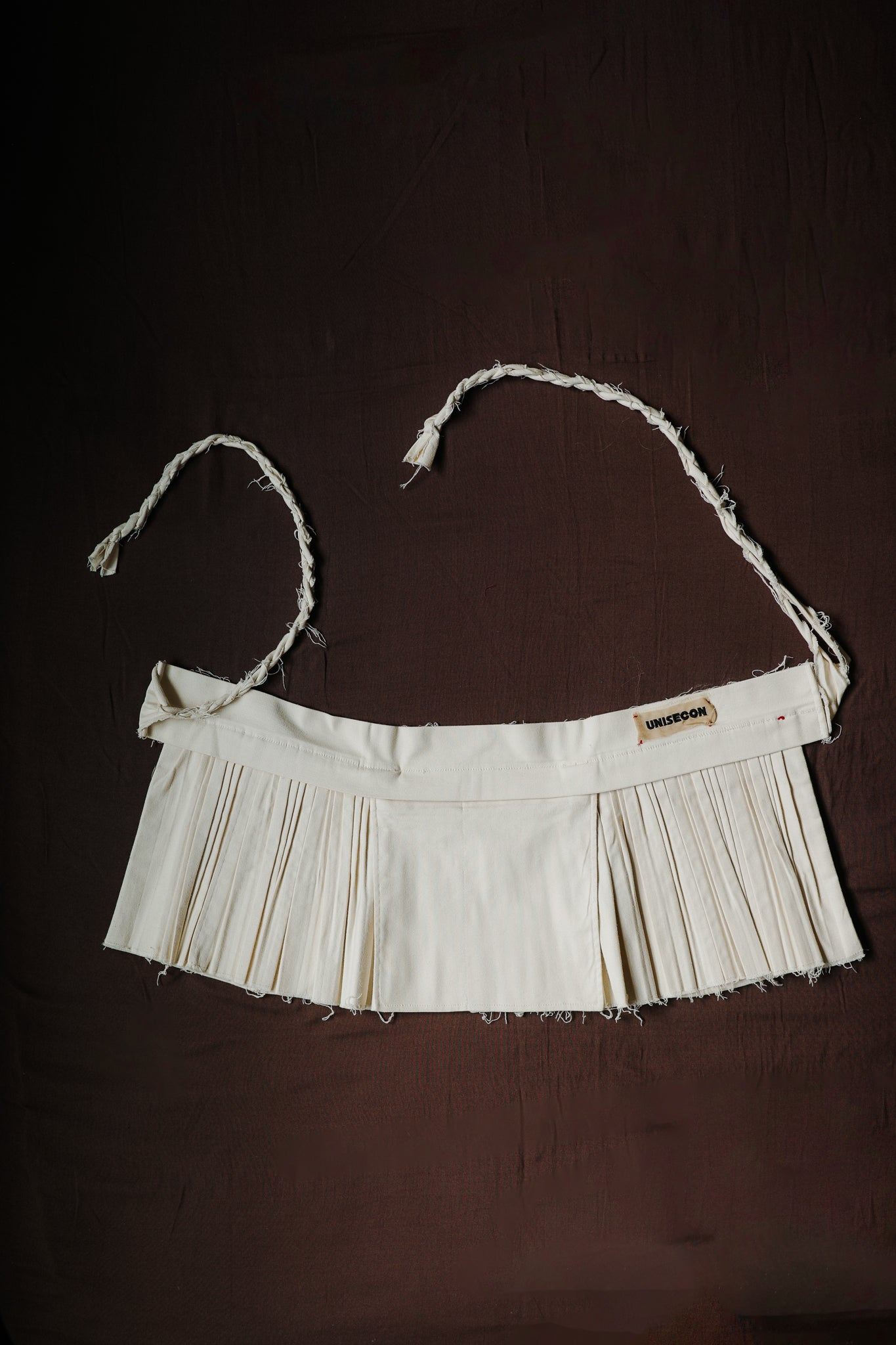 Cream-colored pleated Goma apron with braided ties and raw edges