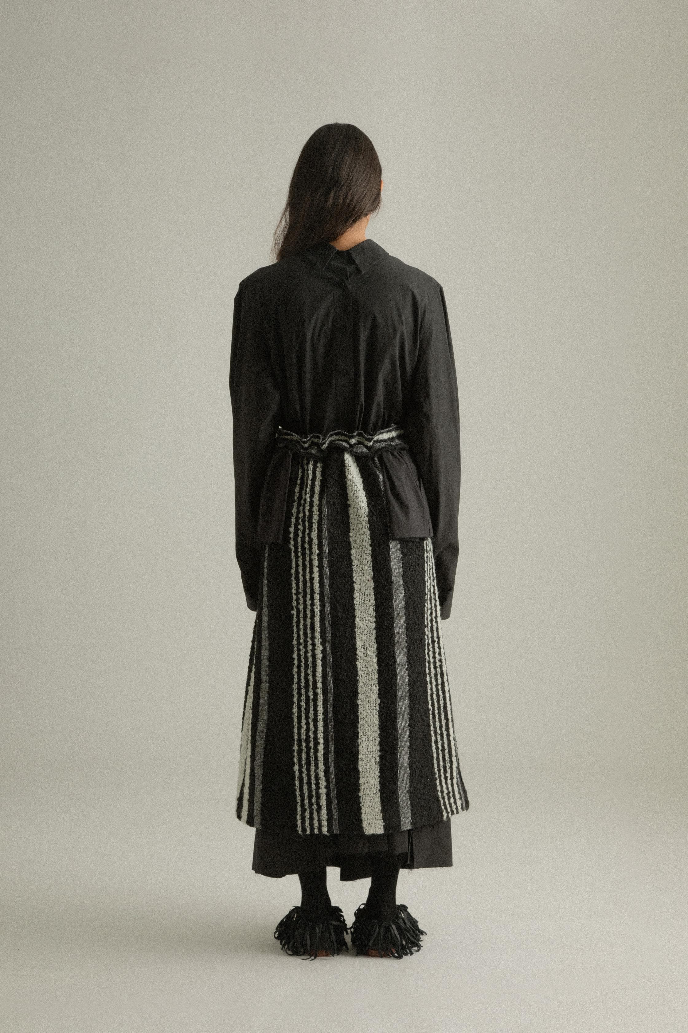 A model wearing the Bing Top, a structured, long-sleeved top with subtle front pleats and a back-tied design made from 100% organic cotton. The top is paired with the Farah Skirt, offering a cohesive look. The model is shown in front, side, and back views to highlight the top’s intricate details and fit.