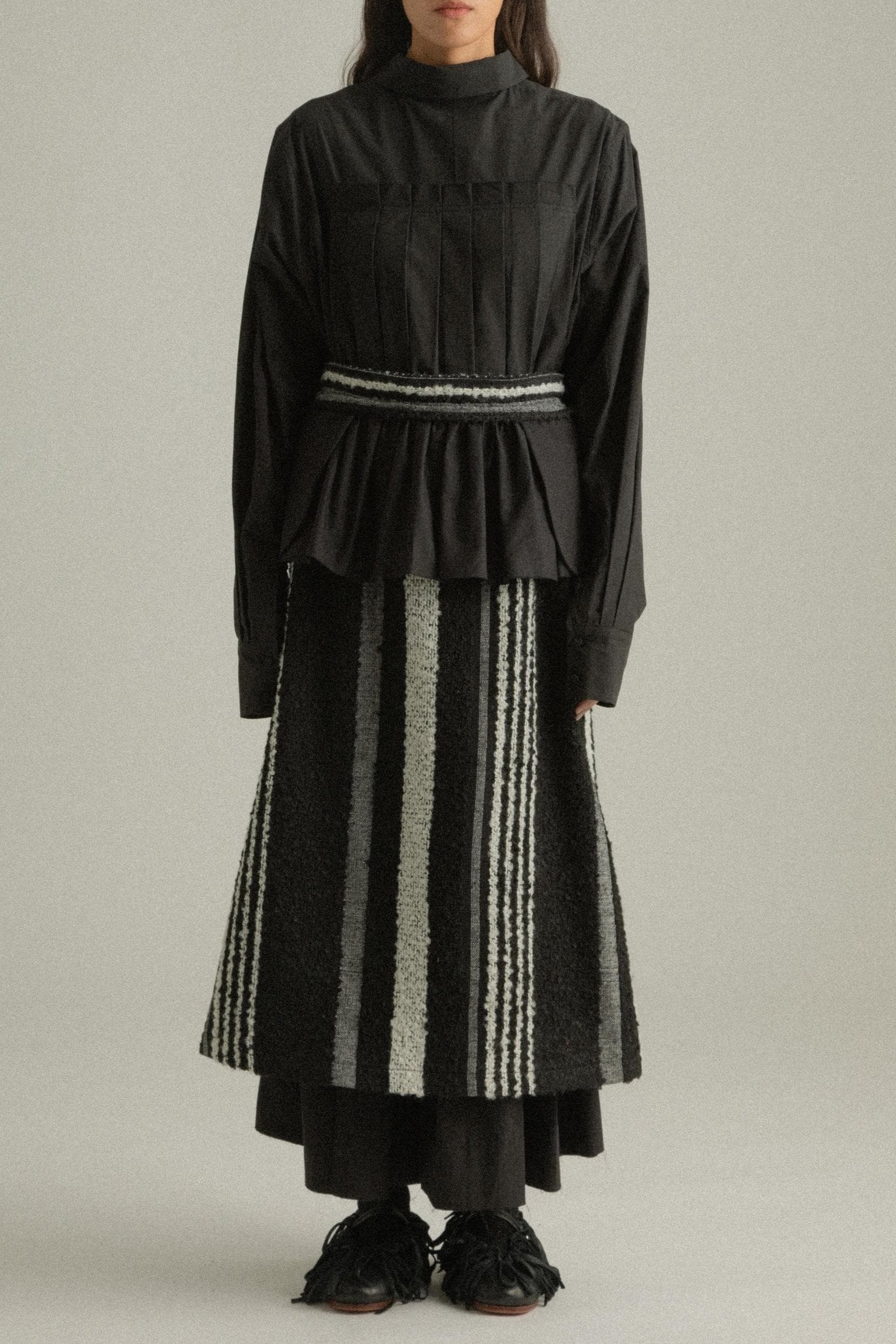 A model wearing the Bing Top, a structured, long-sleeved top with subtle front pleats and a back-tied design made from 100% organic cotton. The top is paired with the Farah Skirt, offering a cohesive look. The model is shown in front, side, and back views to highlight the top’s intricate details and fit.