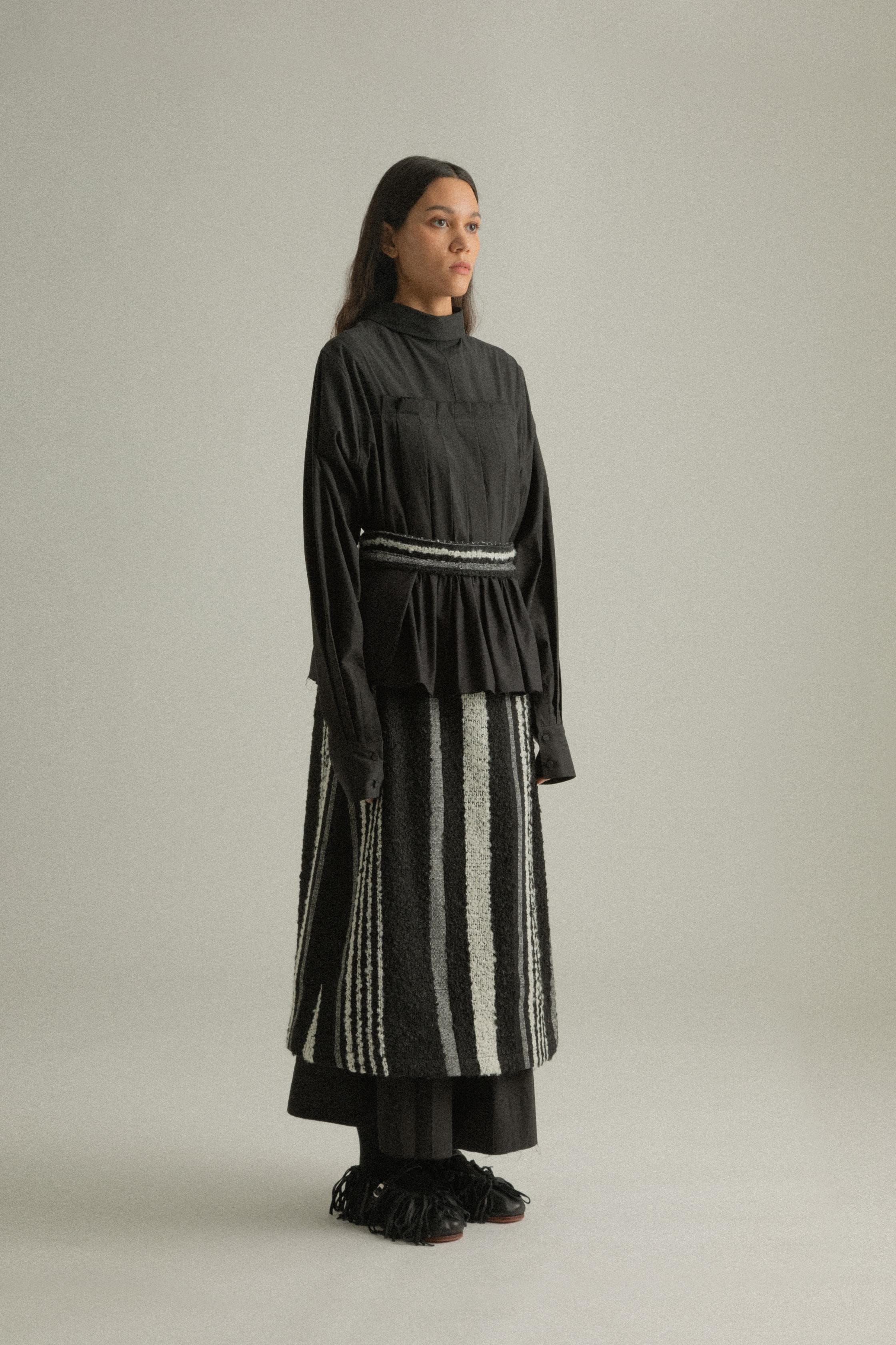 A model wearing the Bing Top, a structured, long-sleeved top with subtle front pleats and a back-tied design made from 100% organic cotton. The top is paired with the Farah Skirt, offering a cohesive look. The model is shown in front, side, and back views to highlight the top’s intricate details and fit.