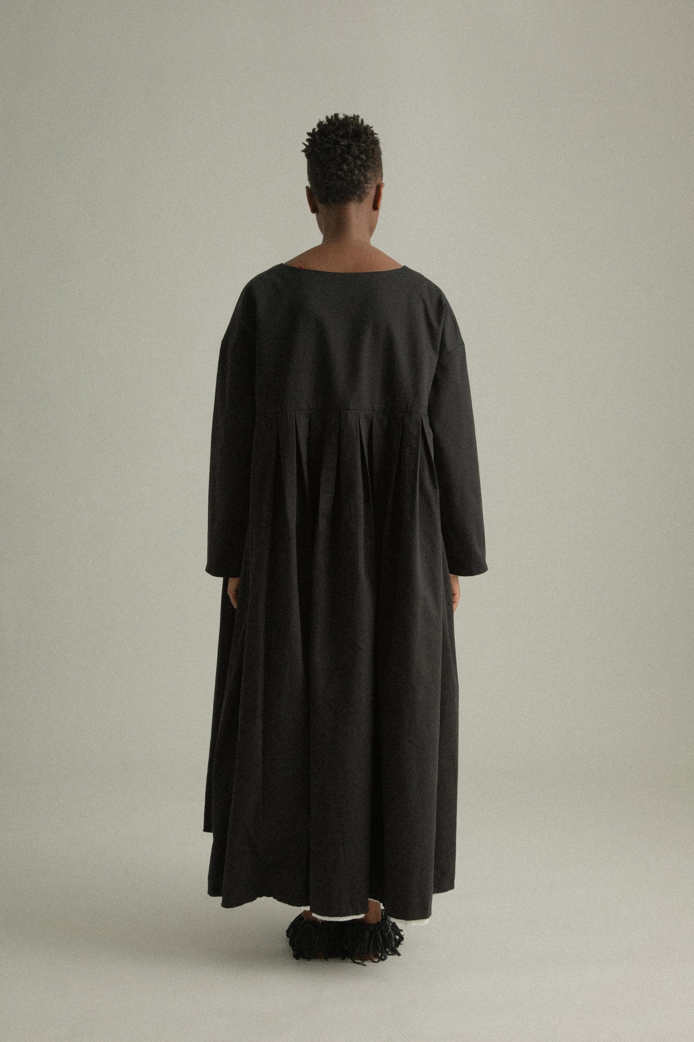 A model wearing the Arya Dress, a loose-fitting, ankle-length dress made of 100% organic cotton. The dress features an asymmetrical neckline, subtle pleating, and a relaxed silhouette. It is designed for reversible wear and has side pockets. The inner white lining is slightly visible at the hem, adding a minimalist contrast to the black fabric. The model is shown in front, side, and back views to highlight the dress’s design and fit.