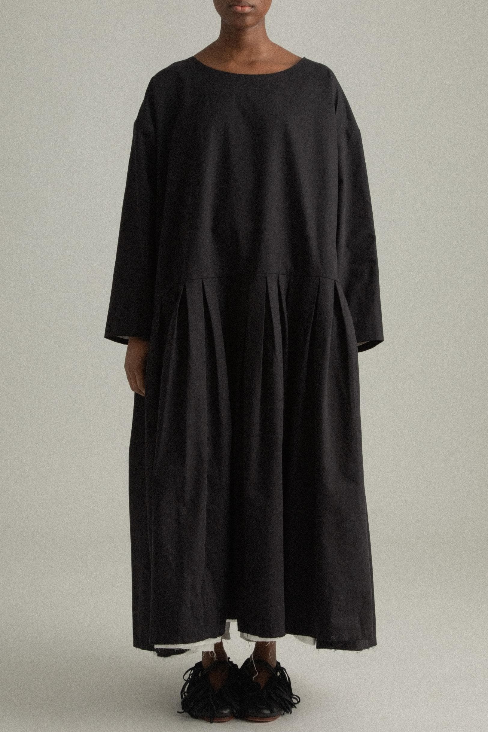 A model wearing the Arya Dress, a loose-fitting, ankle-length dress made of 100% organic cotton. The dress features an asymmetrical neckline, subtle pleating, and a relaxed silhouette. It is designed for reversible wear and has side pockets. The inner white lining is slightly visible at the hem, adding a minimalist contrast to the black fabric. The model is shown in front, side, and back views to highlight the dress’s design and fit.