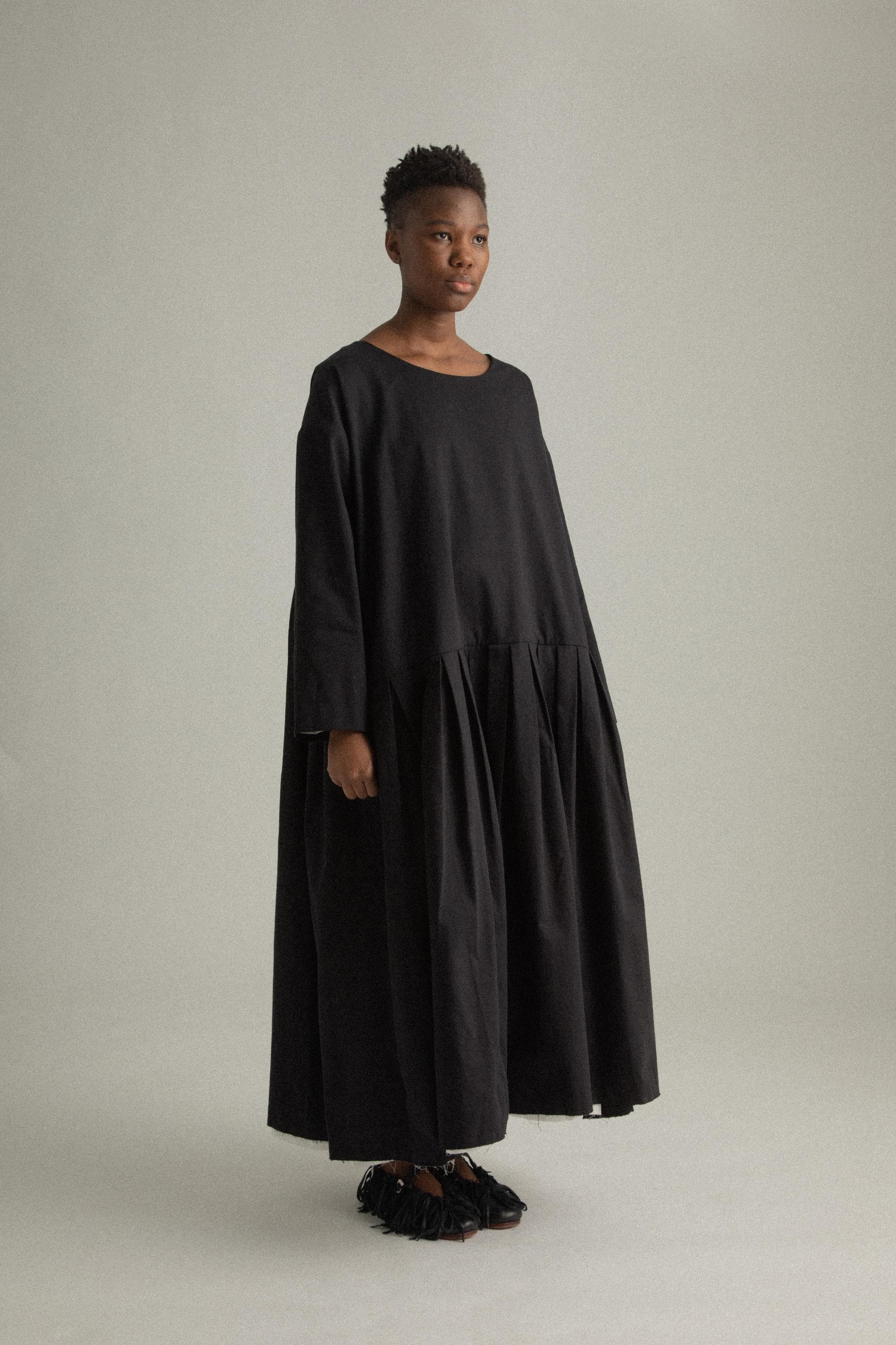 A model wearing the Arya Dress, a loose-fitting, ankle-length dress made of 100% organic cotton. The dress features an asymmetrical neckline, subtle pleating, and a relaxed silhouette. It is designed for reversible wear and has side pockets. The inner white lining is slightly visible at the hem, adding a minimalist contrast to the black fabric. The model is shown in front, side, and back views to highlight the dress’s design and fit.