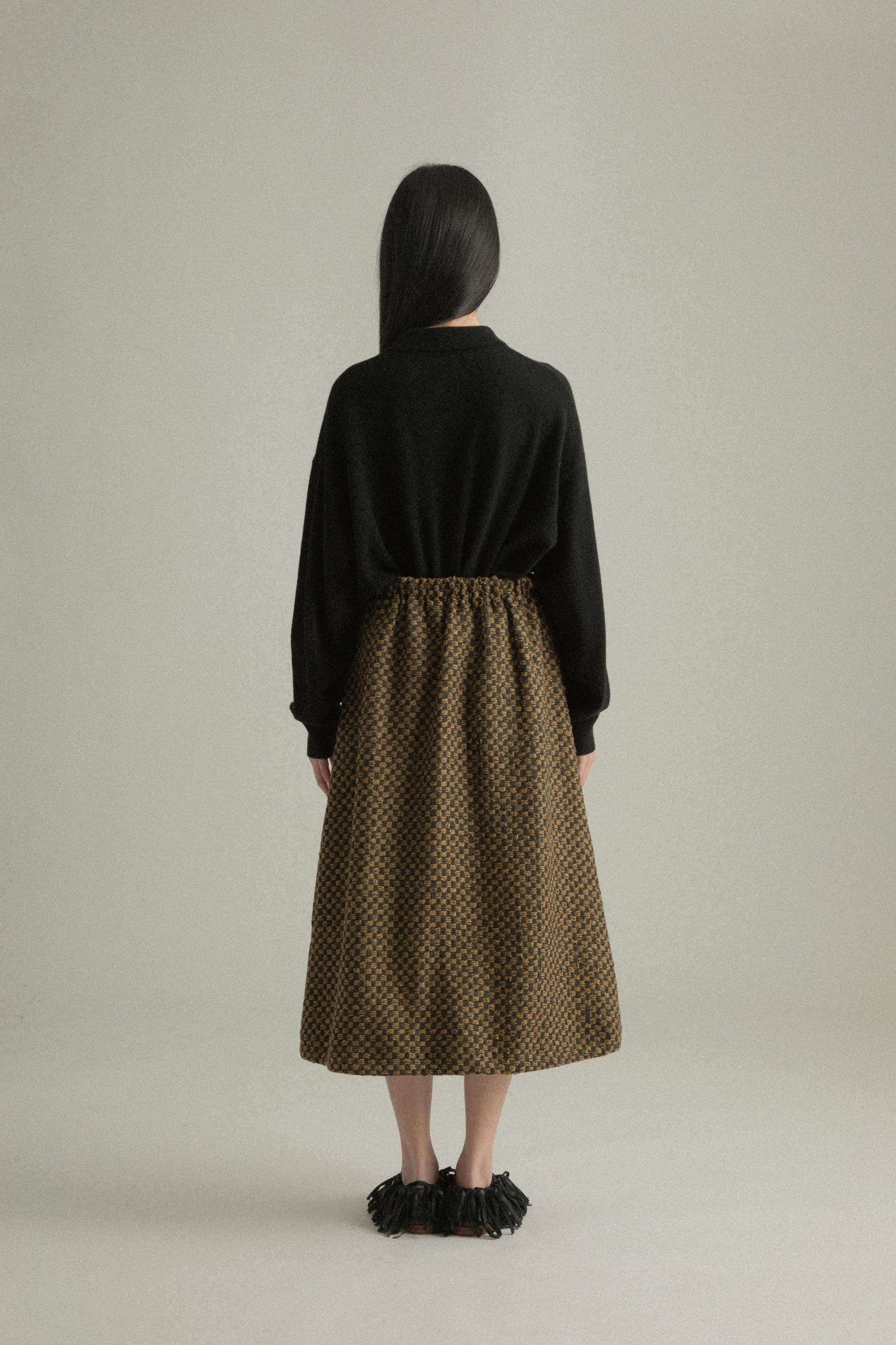 A model wearing the Lulu Skirt, featuring a classic A-line silhouette crafted from a blend of wool and cotton with a jacquard pattern. The skirt showcases a checkered design in black and golden hues. The model is shown in front, side, and back views.