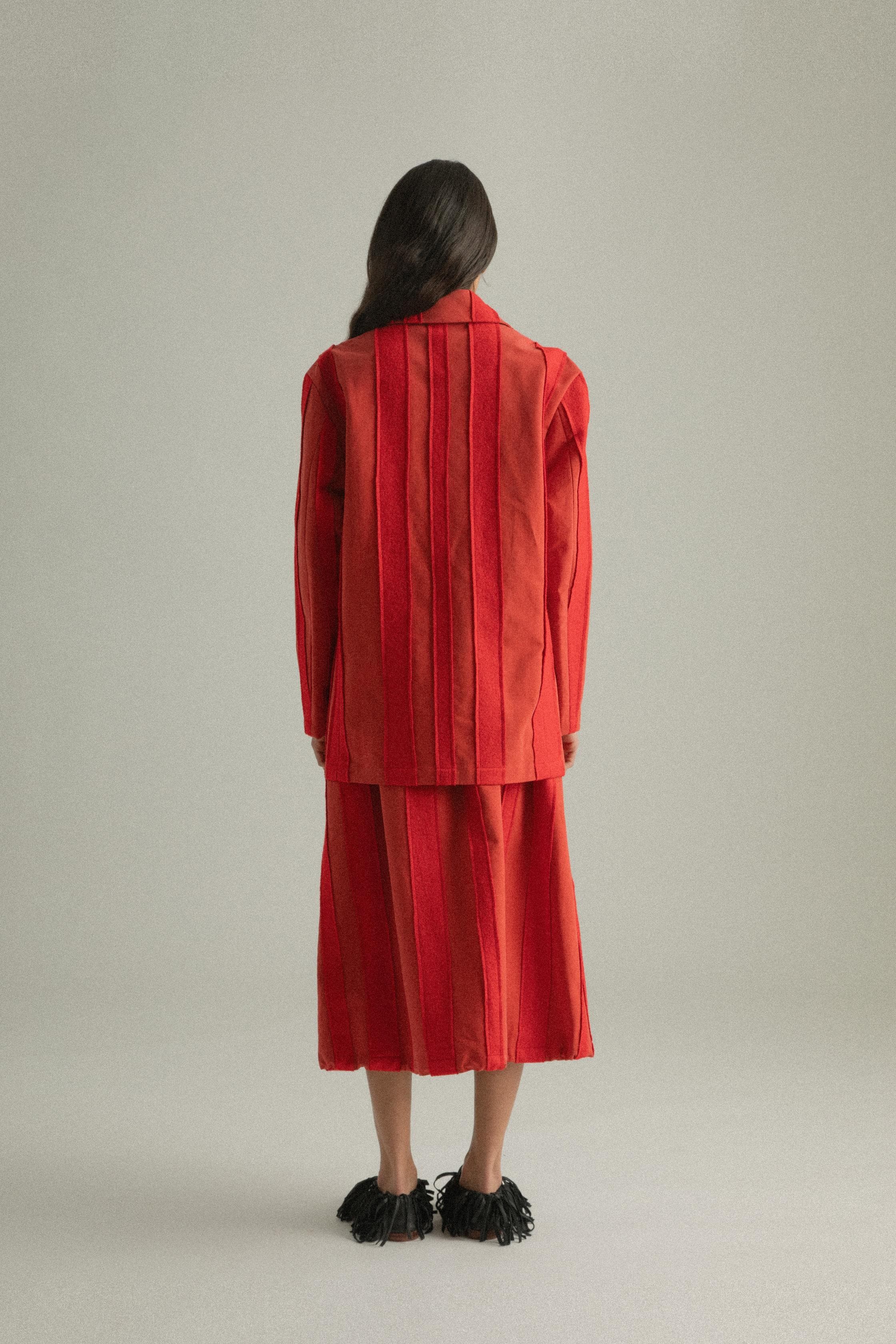 A model wearing the Nicole Jacket in a bold red hue, crafted from 100% cotton with intricate hand-stitching. The jacket features a round collar and single-button closure, styled with the Lulu Skirt. The model is shown in front, side, and back views.