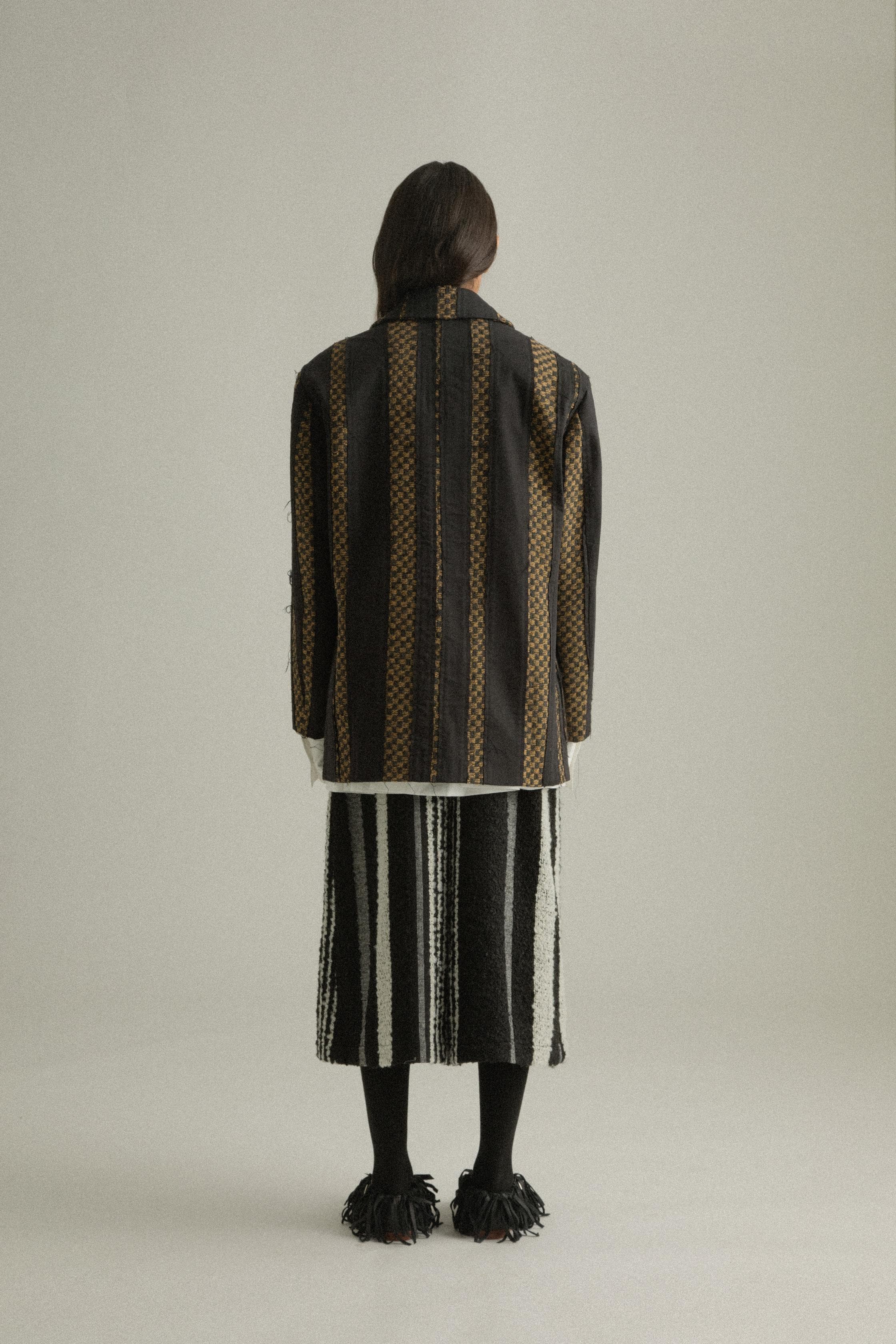 A model wearing the Nicole Jacket, featuring a classic round collar and single-button closure. The jacket is crafted from handwoven fabric in a blend of wool, cotton, and polyester with black and gold hues. Styled with DI Pants for a cohesive look. The model is shown in front, side, and back views.