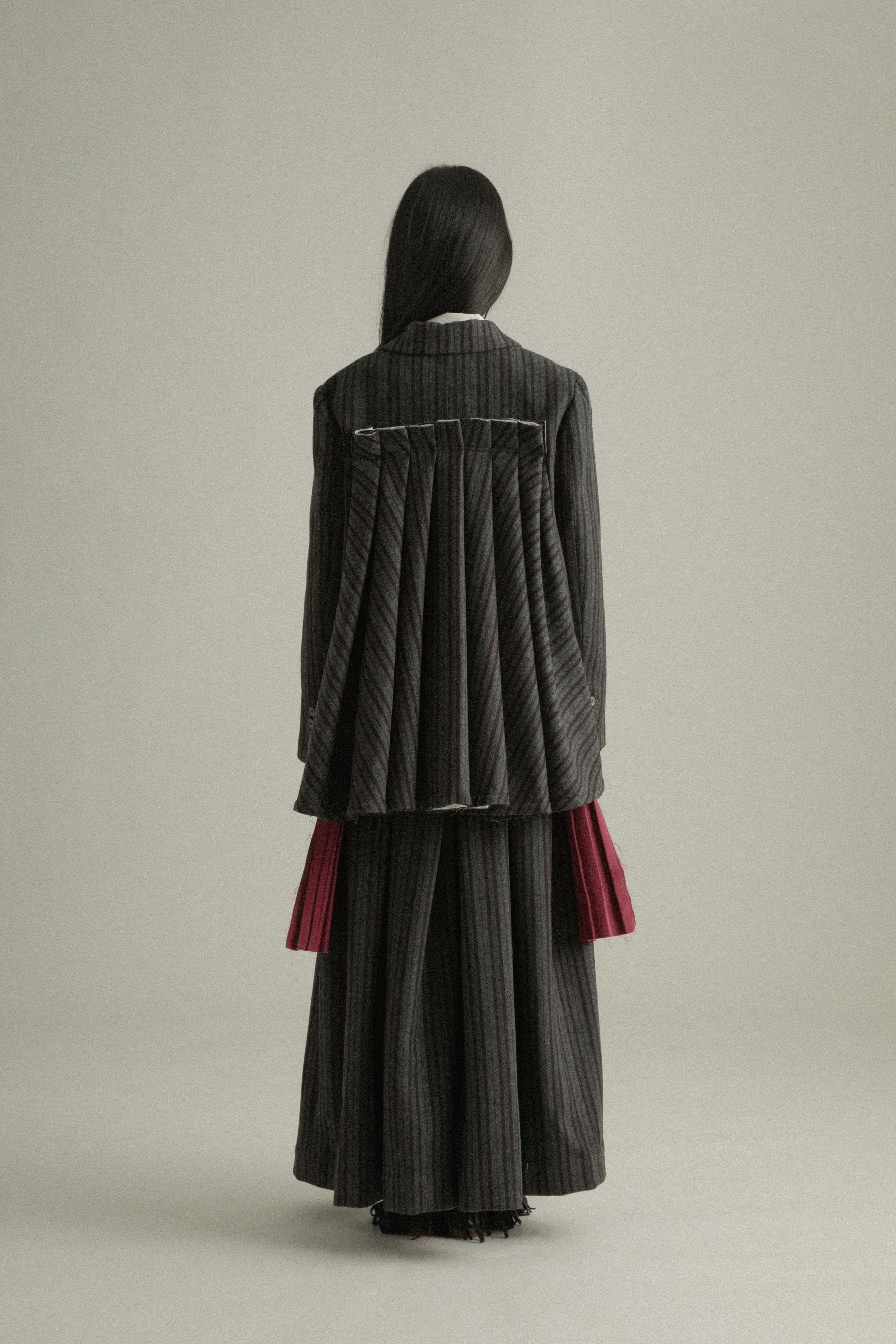 A model wearing the Nina Jacket, crafted from a wool and polyester blend with a dark grey pinstripe pattern and back pleated design. Styled with the Zuri Apron for a playful and sophisticated look. The model is shown in front, side, and back views.