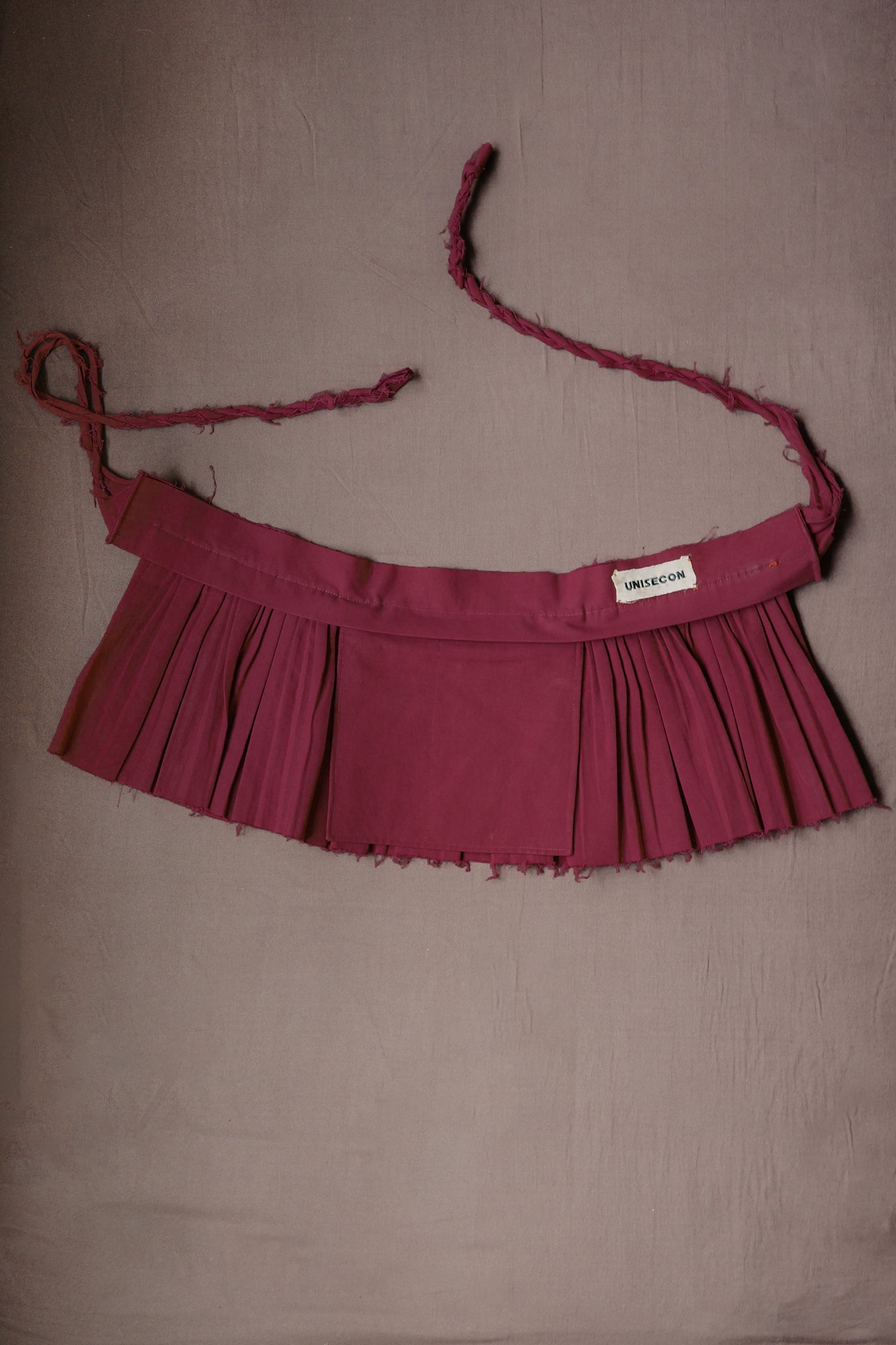 Deep purple pleated Goma apron with structured waistband and braided ties.