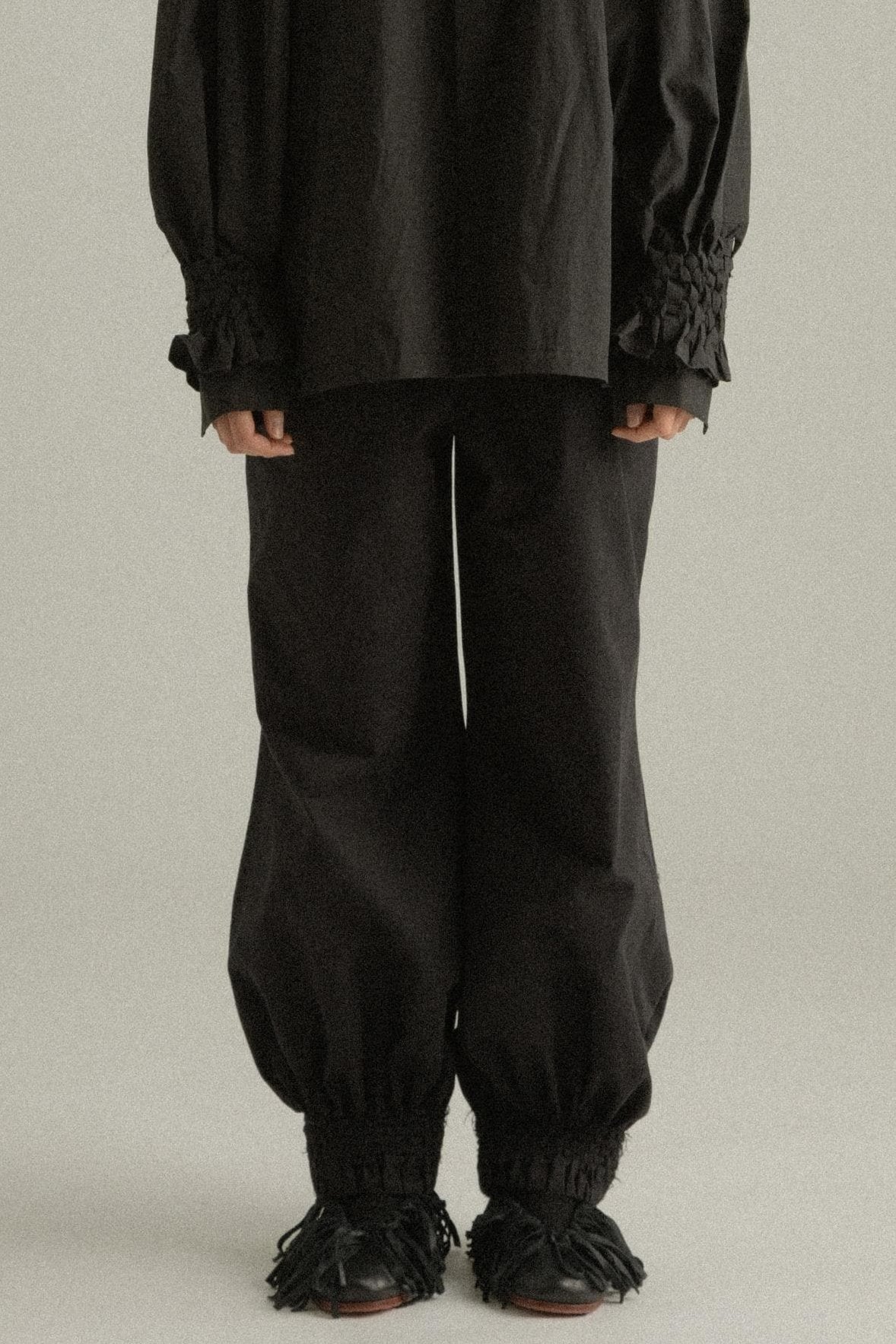 A model wearing the Qira Pants, made from organic cotton with a voluminous silhouette. The pants feature handwoven detailing at the cuffs, an elasticated waistband, and are styled with the Elias Top. The model is shown in front, side, and back views.