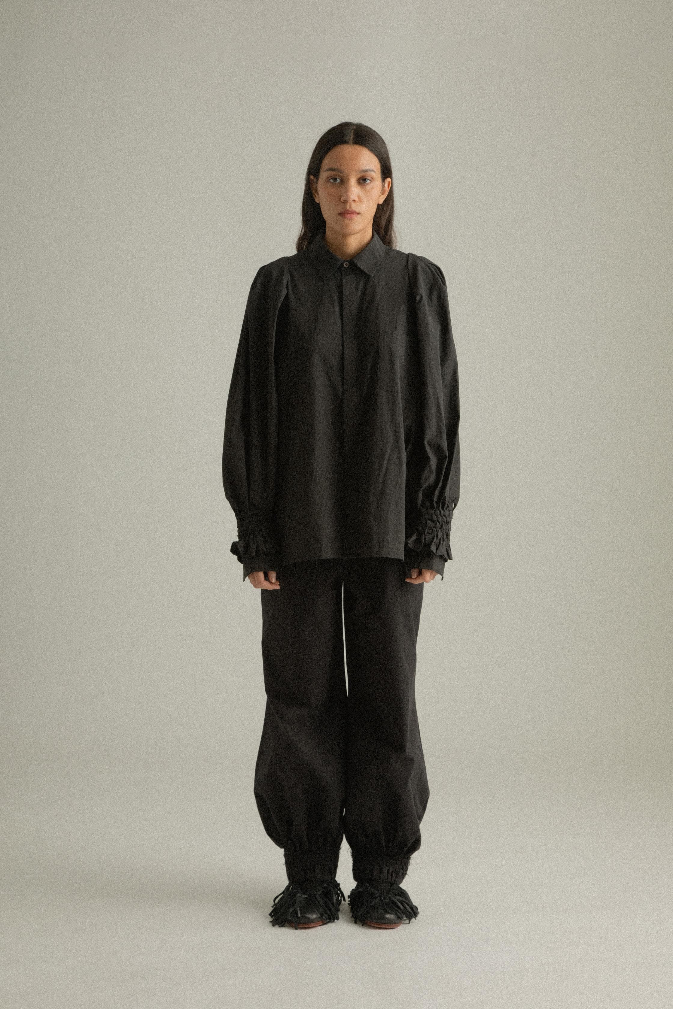 A model wearing the Qira Pants, made from organic cotton with a voluminous silhouette. The pants feature handwoven detailing at the cuffs, an elasticated waistband, and are styled with the Elias Top. The model is shown in front, side, and back views.