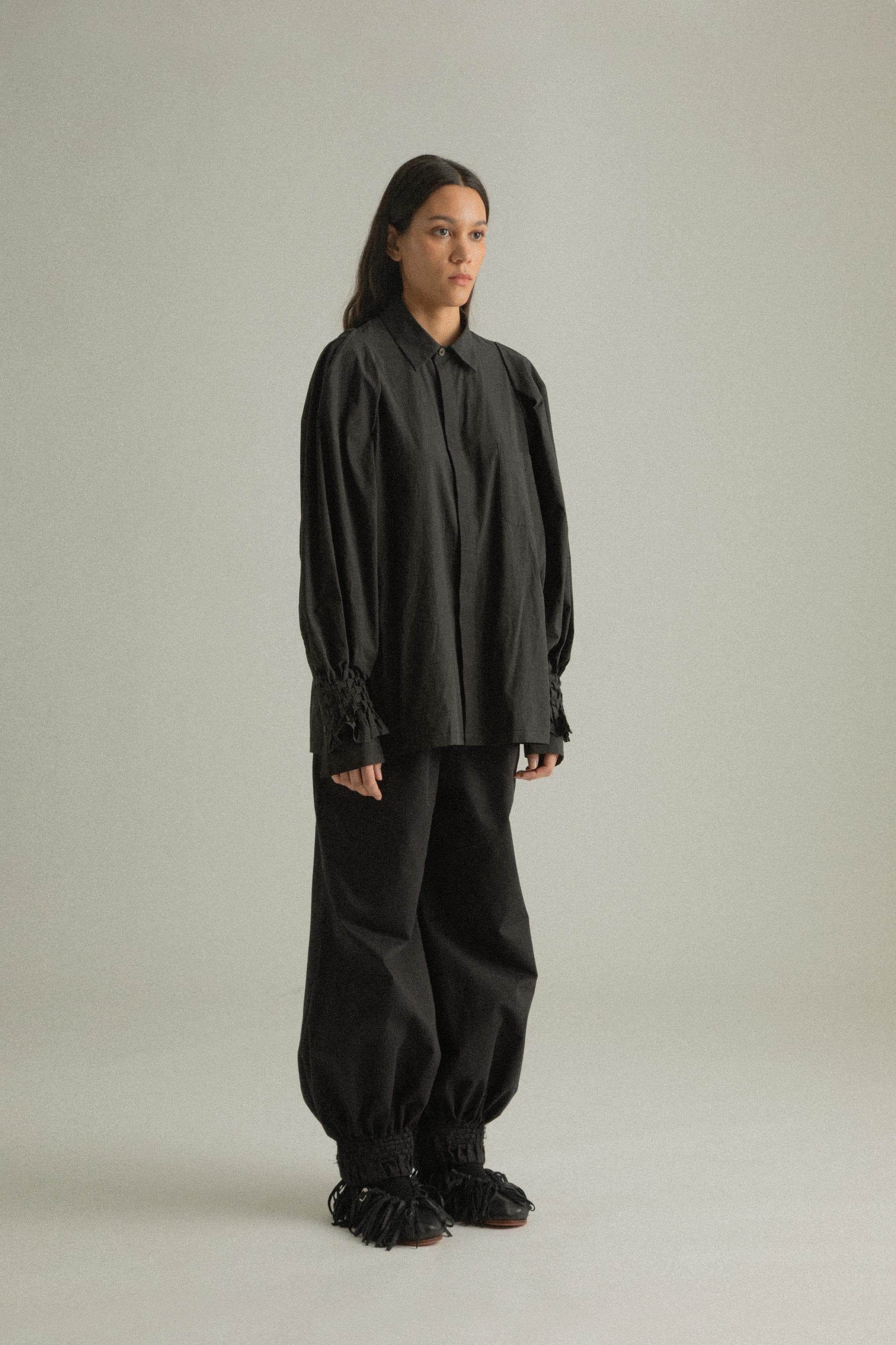 A model wearing the Qira Pants, made from organic cotton with a voluminous silhouette. The pants feature handwoven detailing at the cuffs, an elasticated waistband, and are styled with the Elias Top. The model is shown in front, side, and back views.