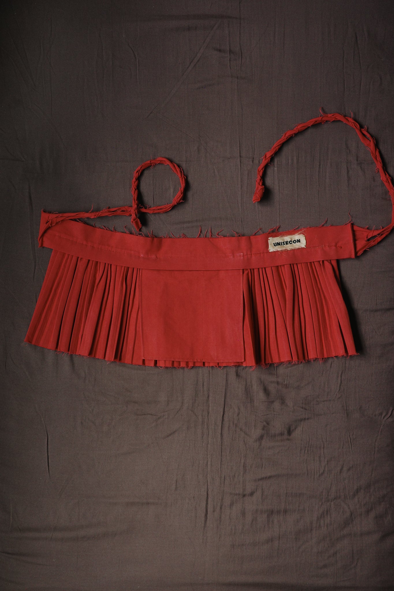 Red pleated Goma apron with hand-braided ties and raw edge details.