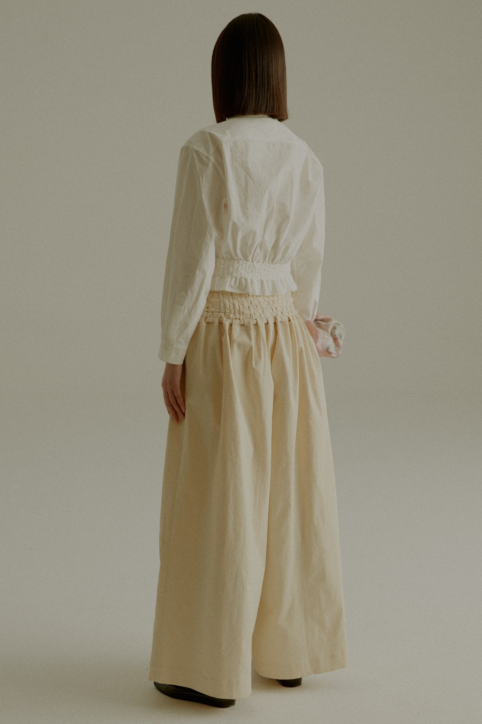 A model wearing the Rose Shirt, a cropped shirt made from 100% organic cotton with handwoven detailing at the waist. Styled with the Sisa Pants. The model is shown in front, side, and back views.