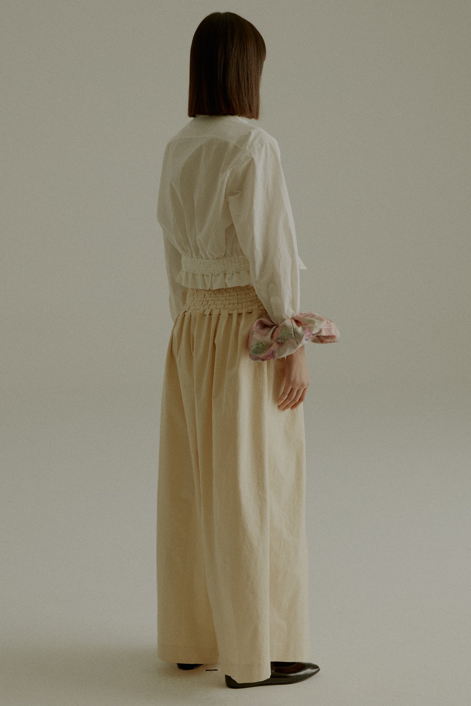 A model wearing the Rose Shirt, a cropped shirt made from 100% organic cotton with handwoven detailing at the waist. Styled with the Sisa Pants. The model is shown in front, side, and back views.