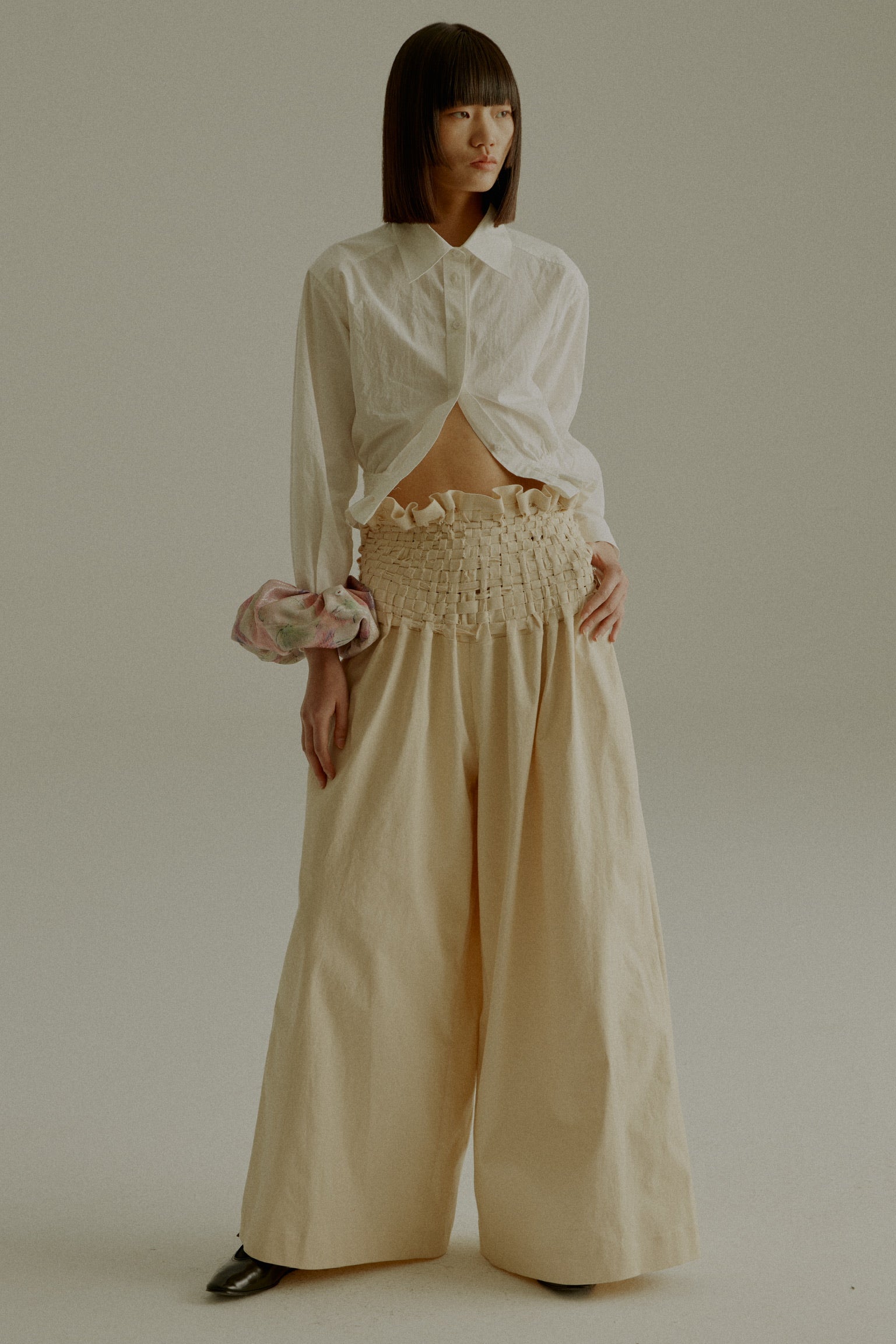 A model wearing the Rose Shirt, a cropped shirt made from 100% organic cotton with handwoven detailing at the waist. Styled with the Sisa Pants. The model is shown in front, side, and back views.