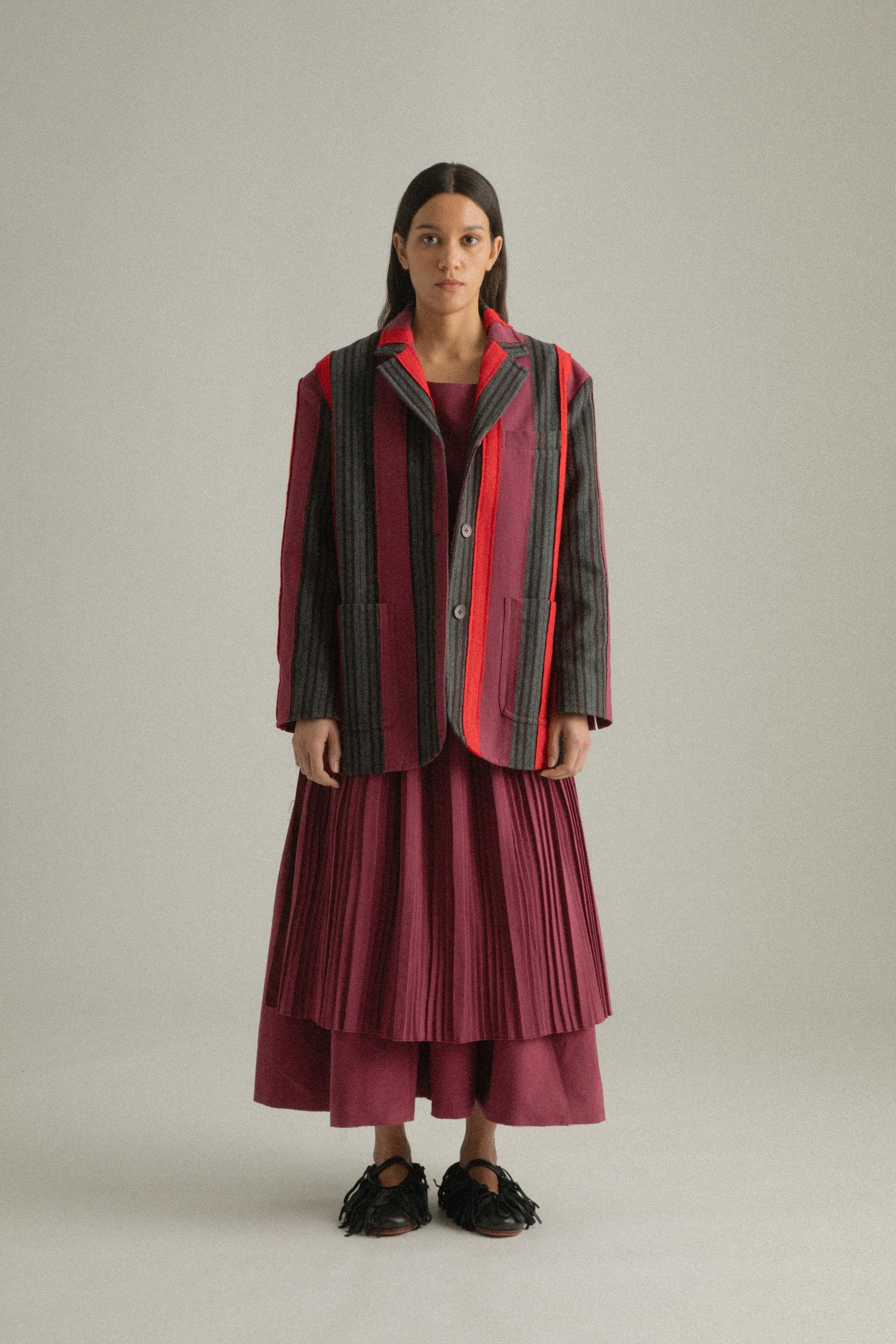 A model wearing the Thais Jacket, featuring a handwoven patchwork design in wool and cotton blends with bold colors. The jacket has a three-button closure and is styled with the Arya Dress. The model is shown in front, side, and back views.