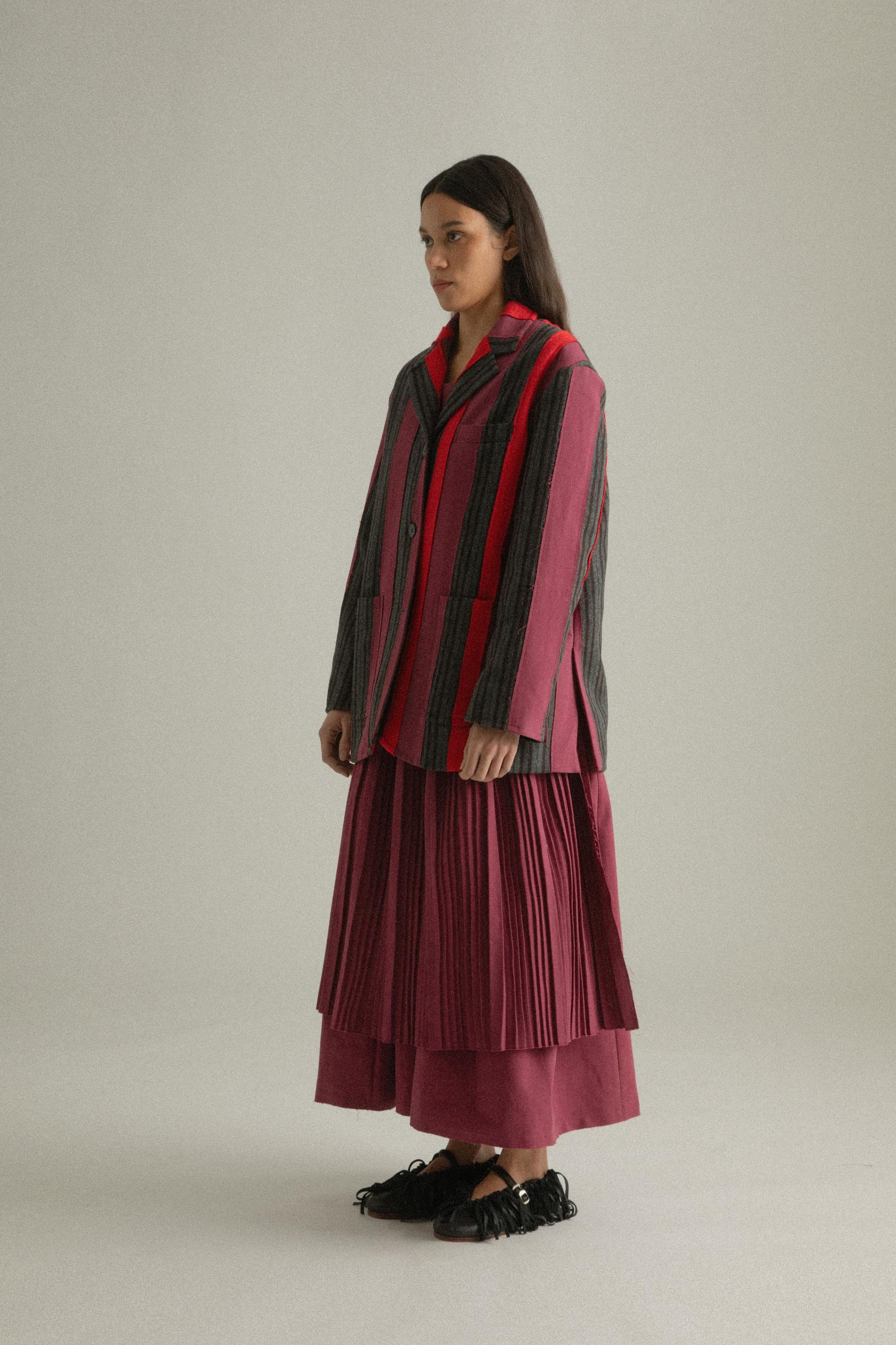 A model wearing the Thais Jacket, featuring a handwoven patchwork design in wool and cotton blends with bold colors. The jacket has a three-button closure and is styled with the Arya Dress. The model is shown in front, side, and back views.